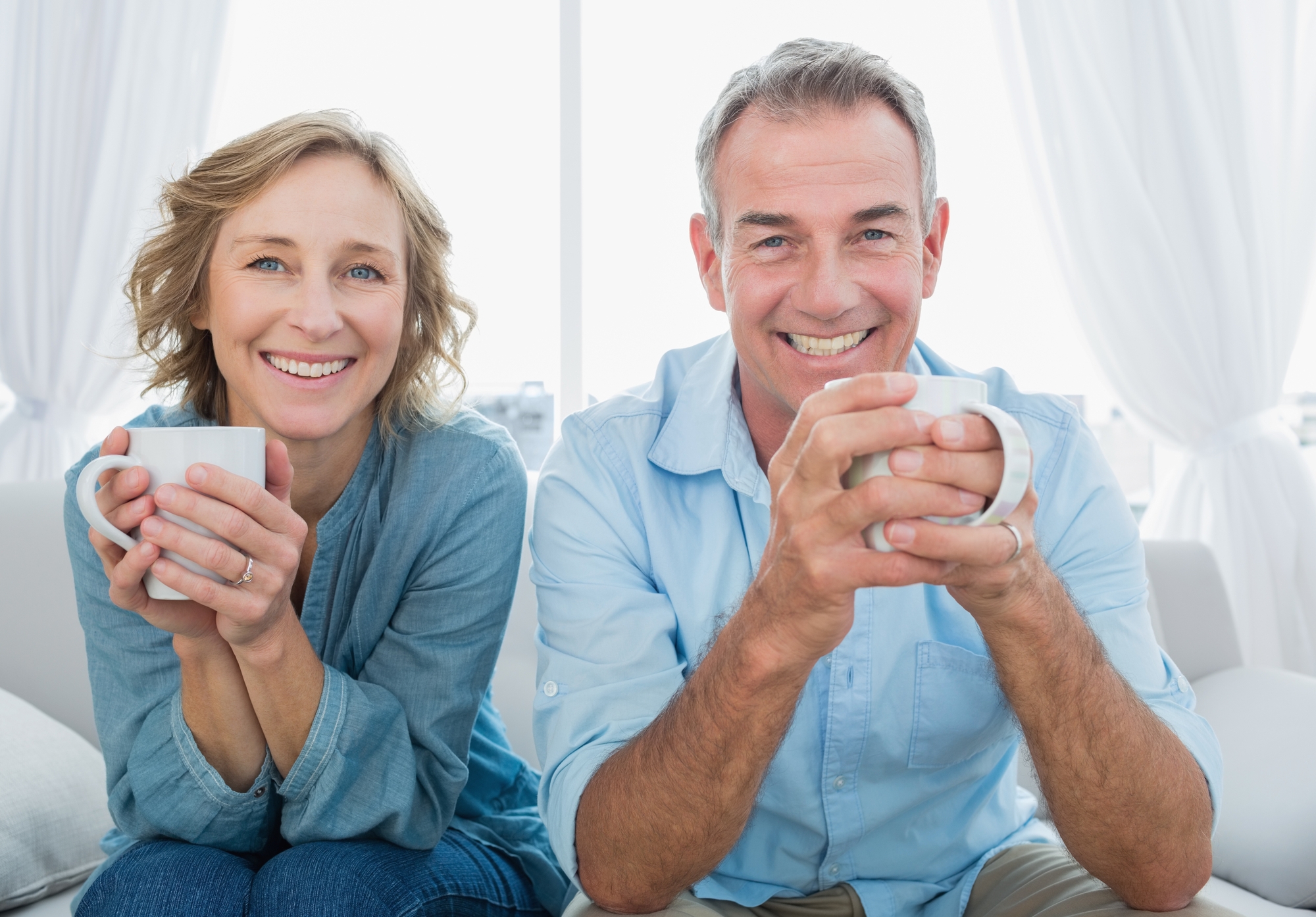 Smiling middle aged couple sitting on the couch having coffee looking at camera at home in the living room